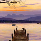 Lake District art print A Perfect Evening Windermere by Jeff Sudders depicting a yellow and orange sunset over the jetty and lake with mountains in shades of purple.