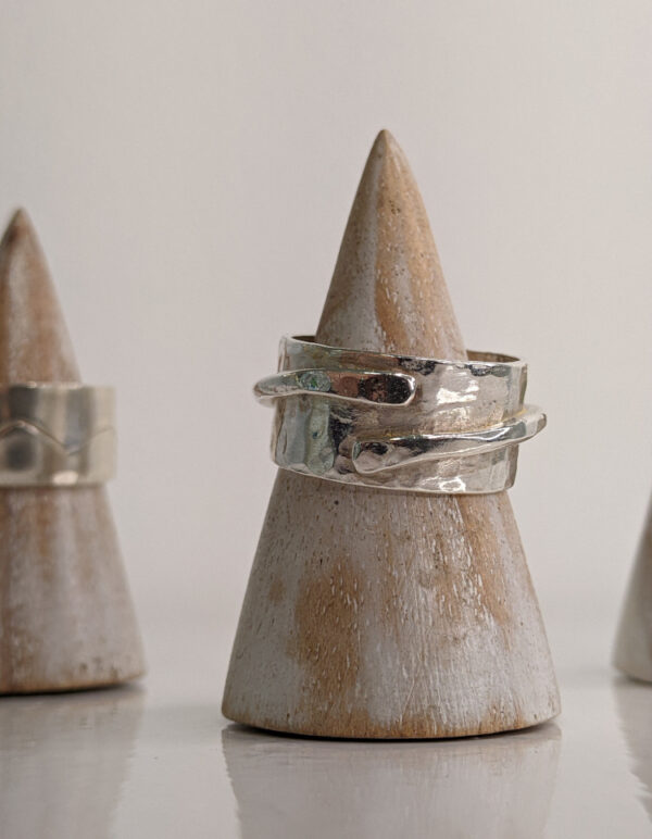 A chunky silver ring with a thin band detail layered over a hammered surface. Approximately 0.75” in diameter. Displayed on a wooden ring stand.