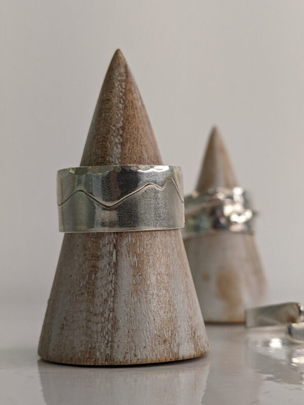 A chunky silver ring featuring a mountain range silhouette wrapping around its band and a hammered texture edge. Approximately 0.75” in diameter.