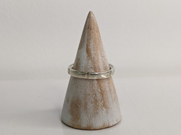 A thin silver ring with small indents across the band. Approximately 0.7” in diameter..Handmade by Helen Wiseman. Displayed on a wooden ring stand.