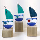 Boats on the block handmade fused glass by Mother and Daughter glass. Blue boats with turquoise or blue sails with red accents. The boats sit on a solid wooden plinth.