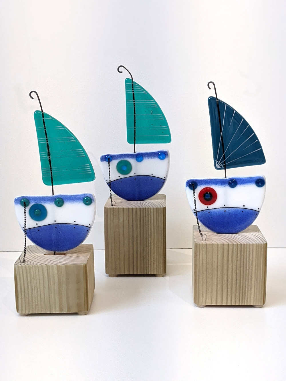 Boats on the block handmade fused glass by Mother and Daughter glass. Blue boats with turquoise or blue sails with red accents. The boats sit on a solid wooden plinth.