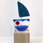 Boats on the block handmade fused glass by Mother and Daughter glass. Blue boat with blue sail and red accents. The boat sits on a solid wooden plinth.