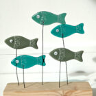 Go With The Flow shoal of fish made using fused glass with hand-painted details and googly eyes. Perched on wires above a wooden plinth swimming along together. Shades of green glass used.