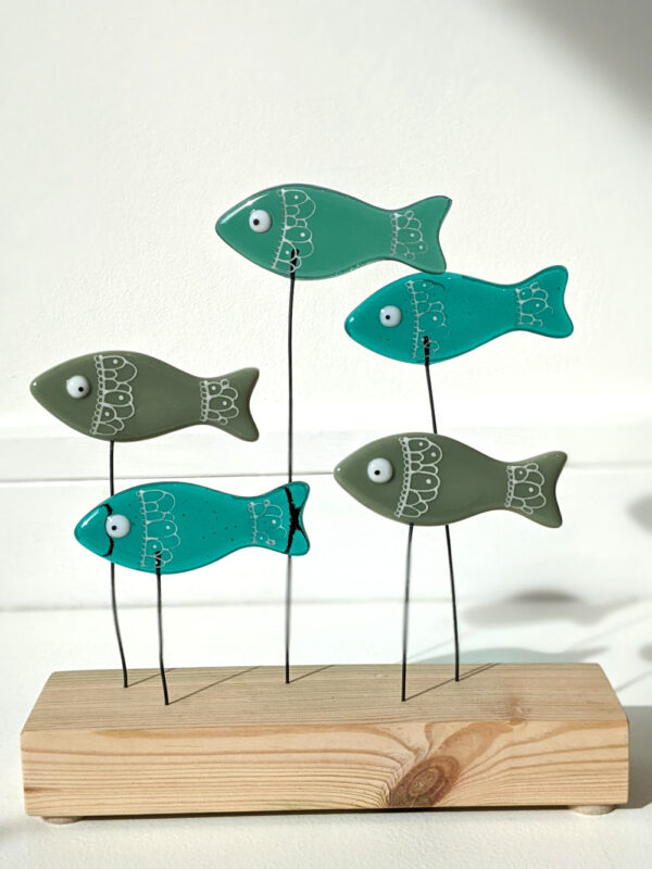 Go With The Flow shoal of fish made using fused glass with hand-painted details and googly eyes. Perched on wires above a wooden plinth swimming along together. Shades of green glass used.