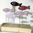 Go With The Flow shoal of fish made using fused glass with hand-painted details and googly eyes. Perched on wires above a wooden plinth swimming along together.. Shades of purple glass used.