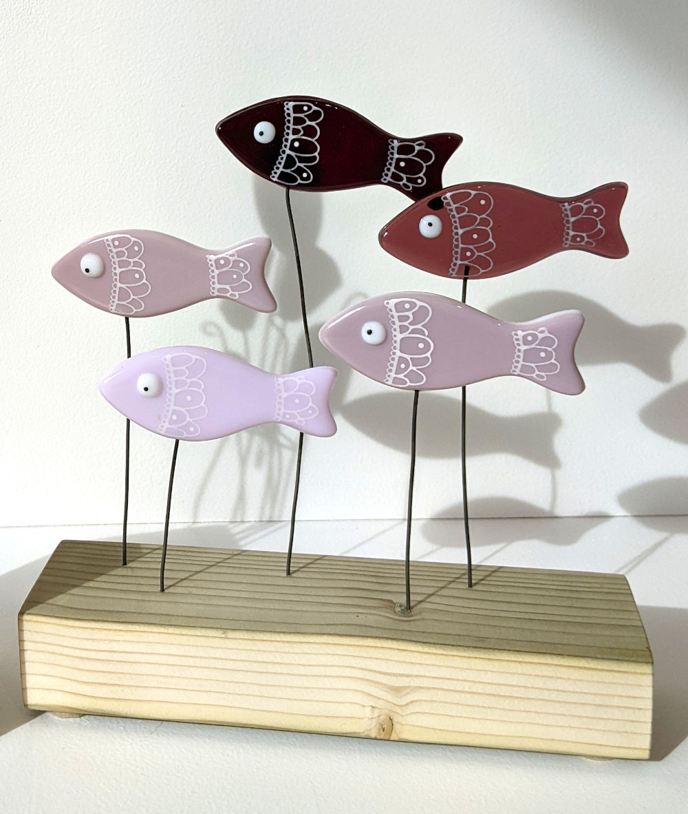Go With The Flow shoal of fish made using fused glass with hand-painted details and googly eyes. Perched on wires above a wooden plinth swimming along together.. Shades of purple glass used.